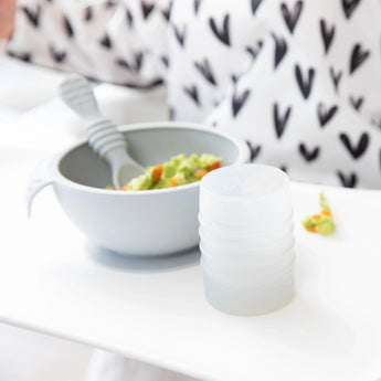 Bumkins Silicone Starter Cup: Gray sits in front of a gray bowl with mashed avocado on a white high chair tray.
