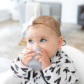 A baby in a white and black bib sipping from a Bumkins Silicone Starter Cup: Gray, while wearing a white bow headband in a high chair.