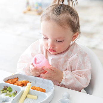 A toddler in a pink bib holds a Bumkins Silicone Starter Cup: Pink, sitting at a table with a gray plate of fruits and snacks.