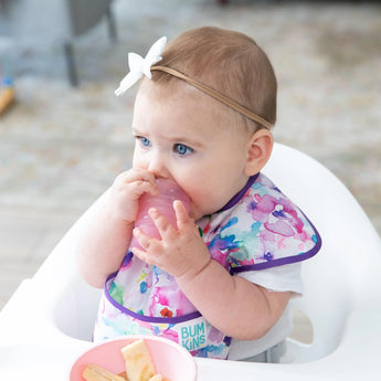 A baby in a bib and bow headband sits in a high chair, holding a Bumkins Silicone Starter Cup: Pink beside snacks.