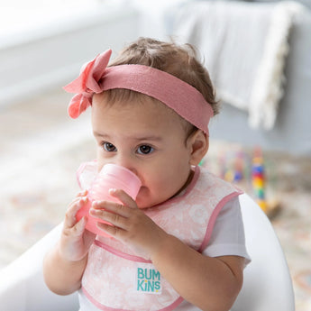 A baby drinks from a Bumkins Silicone Starter Cup: Pink, while wearing a pink headband and bib.