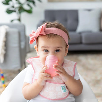 A baby in a pink headband and bib drinks from a Bumkins Silicone Starter Cup: Pink while sitting in a high chair. A gray sofa is behind.