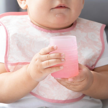 A baby clutches a Bumkins Silicone Starter Cup: Pink while wearing a pink patterned bib.