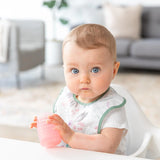A baby with blue eyes sits in a white chair, holding a Bumkins Silicone Starter Cup: Pink with a gray couch in the background.