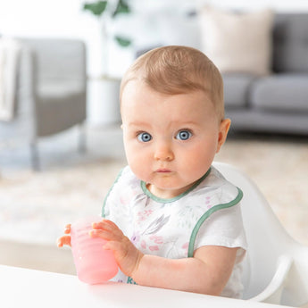 A baby with blue eyes sits in a white chair, holding a Bumkins Silicone Starter Cup: Pink with a gray couch in the background.