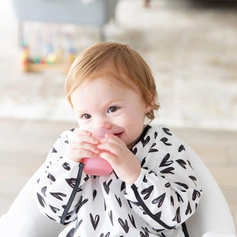 A toddler uses a Bumkins Silicone Starter Cup: Pink while in a high chair, wearing a white bib with black hearts.