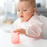 A baby in a patterned bib reaches for a Bumkins Silicone Starter Cup: Pink on a white table.