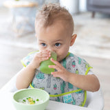 A baby drinks from a Bumkins Silicone Starter Cup: Sage, wearing a cactus bib, with a bowl of food on a high chair tray.
