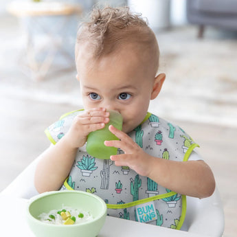 A baby drinks from a Bumkins Silicone Starter Cup: Sage, wearing a cactus bib, with a bowl of food on a high chair tray.