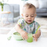 A toddler at a white table holds a Bumkins Silicone Starter Cup: Sage, wearing a cactus bib with a green bowl and spoon beside them.