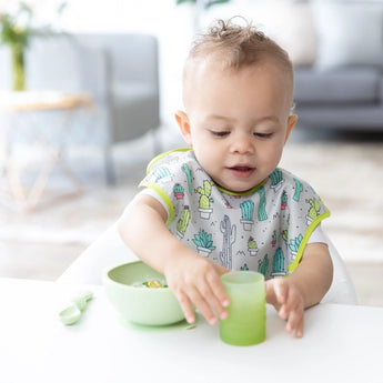 A toddler at a white table holds a Bumkins Silicone Starter Cup: Sage, wearing a cactus bib with a green bowl and spoon beside them.