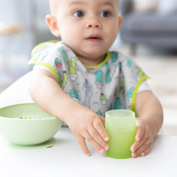 A baby with light hair plays with a Bumkins Silicone Starter Cup: Sage next to a green bowl, wearing a cactus-print bib in a high chair.