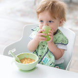 A toddler with brown hair drinks from a Bumkins Silicone Starter Cup: Sage in a high chair, with a green bowl and spoon on the tray.