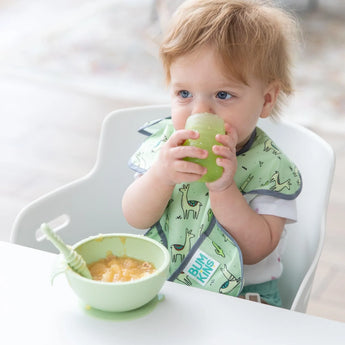 A toddler with brown hair drinks from a Bumkins Silicone Starter Cup: Sage in a high chair, with a green bowl and spoon on the tray.