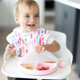 A toddler with a colorful bib opens a pink Bumkins Grip Plate with Silicone Stretch Lid filled with fruits and food.