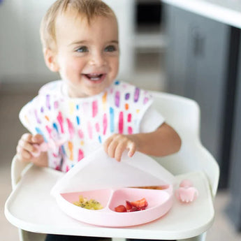 A toddler with a colorful bib opens a pink Bumkins Grip Plate with Silicone Stretch Lid filled with fruits and food.