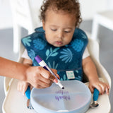 A toddler in a high chair watches an adult write on the Bumkins Silicone Stretch Lid for Grip Dish 3 Section with a purple marker.