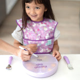 A child wearing a purple bib smiles at Bumkins Grip Plate with Silicone Stretch Lid labeled Madelynes Plate, featuring a fork and spoon.