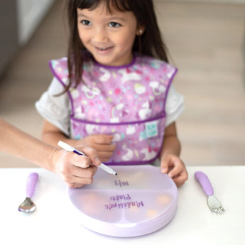 A child wearing a purple bib smiles at Bumkins Grip Plate with Silicone Stretch Lid labeled Madelynes Plate, featuring a fork and spoon.