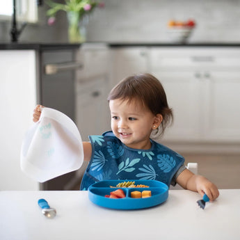 A toddler with a blue bib sits with Bumkins CPSIA compliant dishware, holding a white bib and smiling.