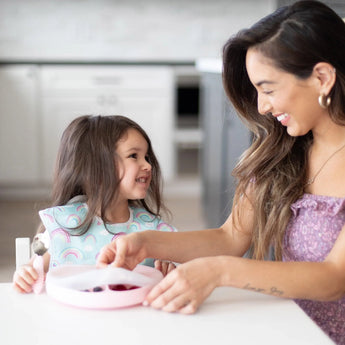 A woman and young girl with Bumkins rainbow bib and silicone dishware share smiles at the table, CPSIA Compliant joy!.