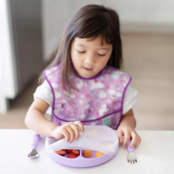 A young child with a purple bib enjoys fruit from a Bumkins silicone dishware set, using a utensil.