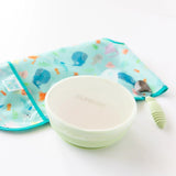 Bumkins Silicone Stretch Lid for Grip Bowl covers a green bowl next to a green spoon and folded ocean-themed bib on a white background.