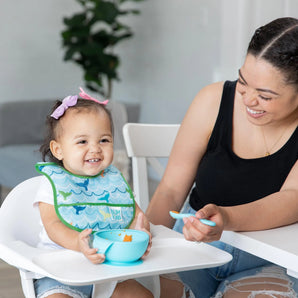 A smiling woman feeds a happy toddler in a high chair wearing a Bumkins Starter Bib 2 Pack: Ocean Life & Whale Tail from a blue bowl.