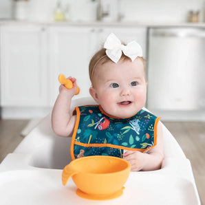 A baby sits in a high chair, wearing a Bumkins Starter Bib 2 Pack: Jungle & Animal Prints and holding an orange spoon near a matching bowl.