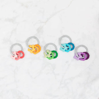 Five teething toys sit on a white marble background, including Bumkins Silicone Teething Charms: Orange.