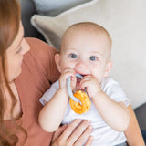 A baby with big blue eyes chews on a Bumkins Silicone Teething Charms: Orange, offering teething relief, held by a red-haired woman.