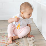 A baby in a white bow headband sits on a rug, holding a Bumkins Silicone Teething Charms: Orange by white shelves.