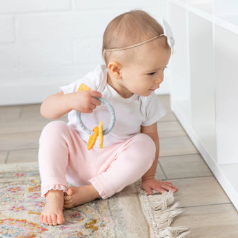 A baby in a white bow headband sits on a rug, holding a Bumkins Silicone Teething Charms: Orange by white shelves.