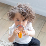 A toddler with curly hair in a white shirt and black pants chews on a Bumkins Silicone Teething Charms: Orange while seated on a wood floor.