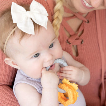 A baby with a white bow chews on a Bumkins Silicone Teething Charms: Orange, held by someone in a peach sweater.