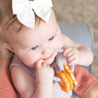 A baby in a purple outfit, wearing a white bow, enjoys teething relief with Bumkins Silicone Teething Charms: Orange on someone's lap.