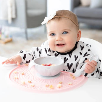 A smiling baby sits at a table in a patterned shirt and headband, with a bowl and spoon on a pink placemat, wearing Bumkins Sleeved Bib: Hearts.