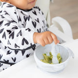 A toddler in a Bumkins Sleeved Bib: Hearts mashes avocado with a spoon in a white bowl.