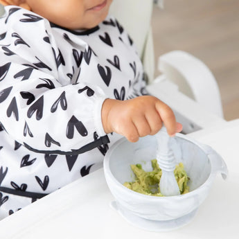 A toddler in a Bumkins Sleeved Bib: Hearts mashes avocado with a spoon in a white bowl.