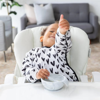 A toddler in a high chair wears a Bumkins Sleeved Bib: Hearts, giving a thumbs-up while holding a spoon by an empty bowl.