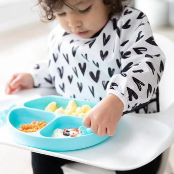 Toddler in Bumkins Sleeved Bib: Hearts, using a blue sectioned plate and spoon to scoop yogurt, with crumb catcher in action.