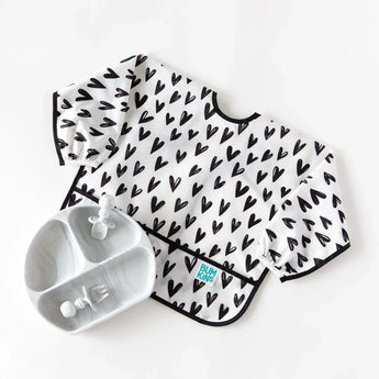 Bumkins Sleeved Bib: Hearts with crumb catcher next to a marbled gray divided plate and utensils on a white background.