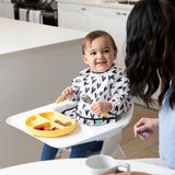 A toddler in a high chair smiles at an adult while using a spoon and eating from a yellow plate, wearing Bumkins Sleeved Bib: Hearts.