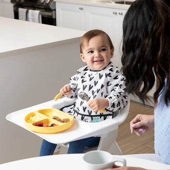 A toddler in a high chair smiles at an adult while using a spoon and eating from a yellow plate, wearing Bumkins Sleeved Bib: Hearts.