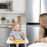 A toddler wearing a Bumkins Sleeved Bib: Hearts and heart-patterned shirt holds a spoon, smiling at an adult in a modern kitchen.