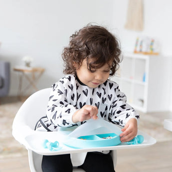 A toddler with curly hair sits in a high chair, wearing a Bumkins Sleeved Bib: Hearts. They focus on eating from a blue divided plate on the tray. The softly lit room, with charming furniture and decor in the background, completes the scene.
