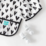 Bumkins Sleeved Bib: Hearts, waterproof with black-and-white pattern. Spoon and fork nearby for mealtime messes.
