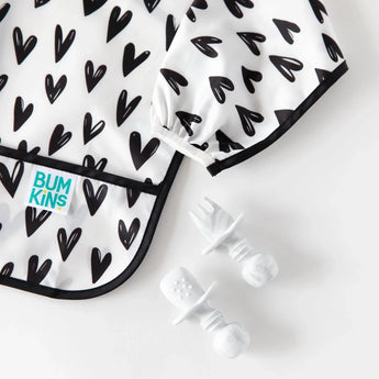 Bumkins Sleeved Bib: Hearts, waterproof with black-and-white pattern. Spoon and fork nearby for mealtime messes.