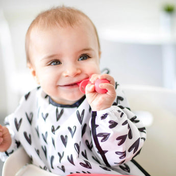 Happy baby in a Bumkins Sleeved Bib: Hearts, holding a pink toy while seated in a high chair. Waterproof and adorable!.