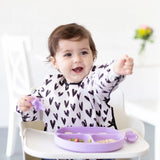 A smiling toddler with dark hair sits in a high chair, wearing a Bumkins Sleeved Bib: Hearts and holding a purple spoon over a neatly arranged purple sectioned plate. The blurred background with chairs and flowers enhances the peaceful atmosphere ideal for easy cleaning.